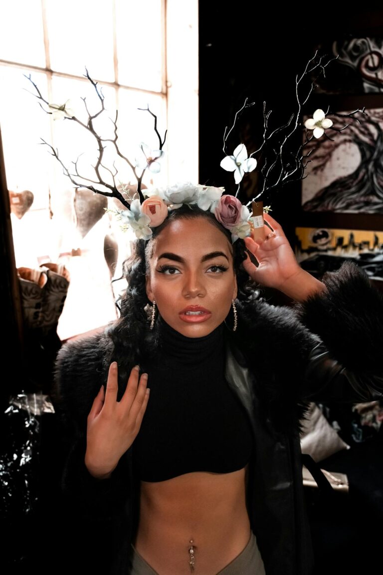 A stylish woman wearing a floral antler headpiece inside a dimly lit artistic room.